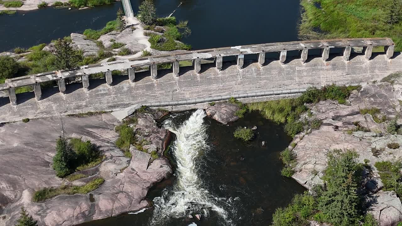 Drone zooming in and orbiting right on the dam wall at Old Pinawa Dam Manitoba, showing water rushing underneath, spillway flow, and concrete hydro structure in summer