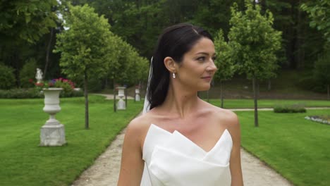 Vista-Frontal-De-La-Hermosa-Novia-Caminando-Con-Una-Sonrisa-Confiada-En-El-Parque-El-Día-De-Su-Boda,-Sosteniendo-Su-Vestido-De-Novia-Y-Mostrando-Piernas-Sexys,-La-Cámara-Sigue-En-El-Día-De-Verano