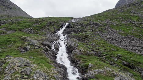 Toma-Aérea-De-Una-Plataforma-Rodante,-Saliendo-De-Un-Río-Estrecho-Y-Caudaloso-Mientras-Desciende-Por-Una-Ladera-Empinada,-Rocosa-Y-Cubierta-De-Musgo,-Trollstigen,-Noruega