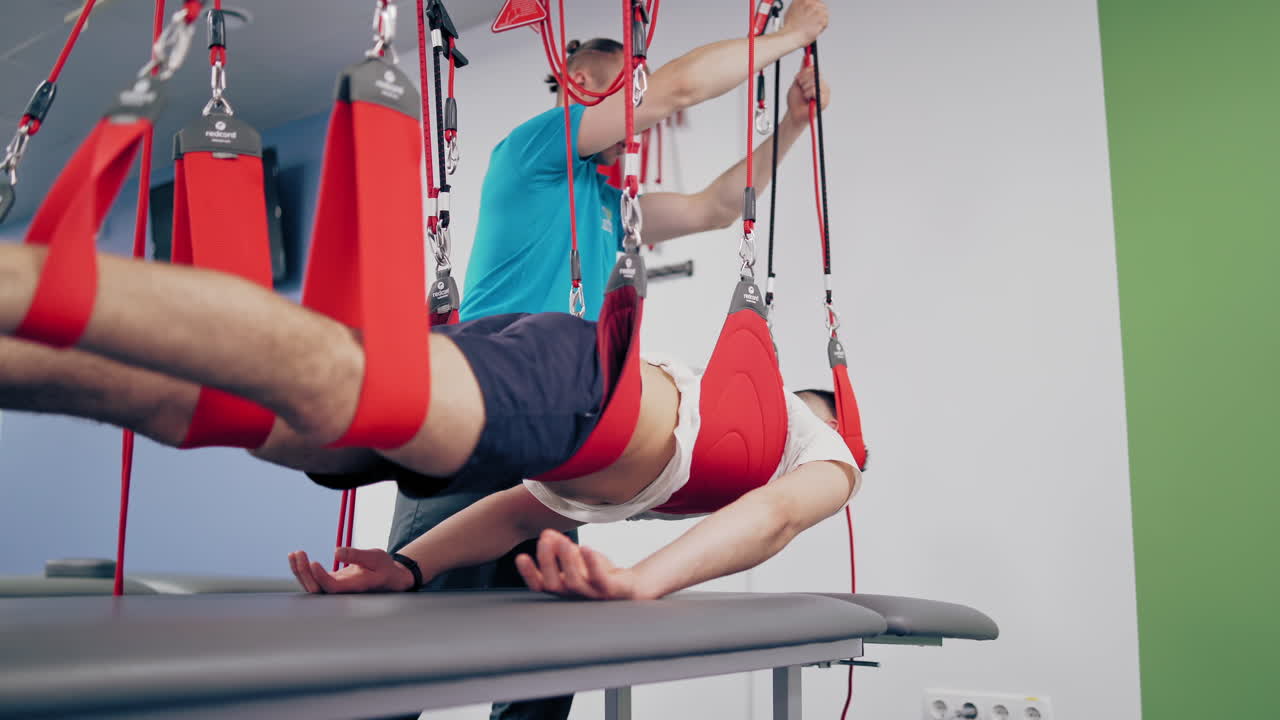 Physiotherapist is rising his client man with a special suspension rope system. Young man is hanging by rope system in a rehabilitation center. Instructor helps his patient to recover.