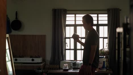 medium side view of young man shaking protein drink by an apartment kitchen window in morning light