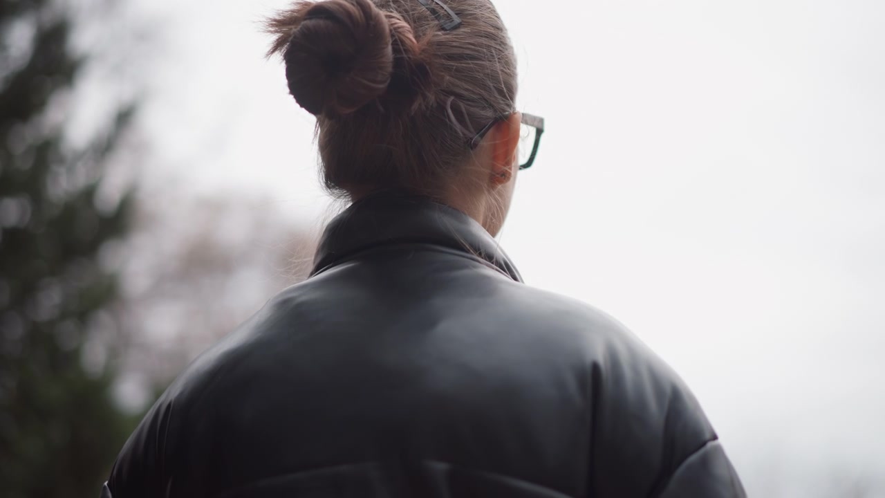 Close up of woman with brown hair in bun wearing glasses and black leather jacket looking into distance. background features blurred trees under overcast sky creating calm outdoor setting