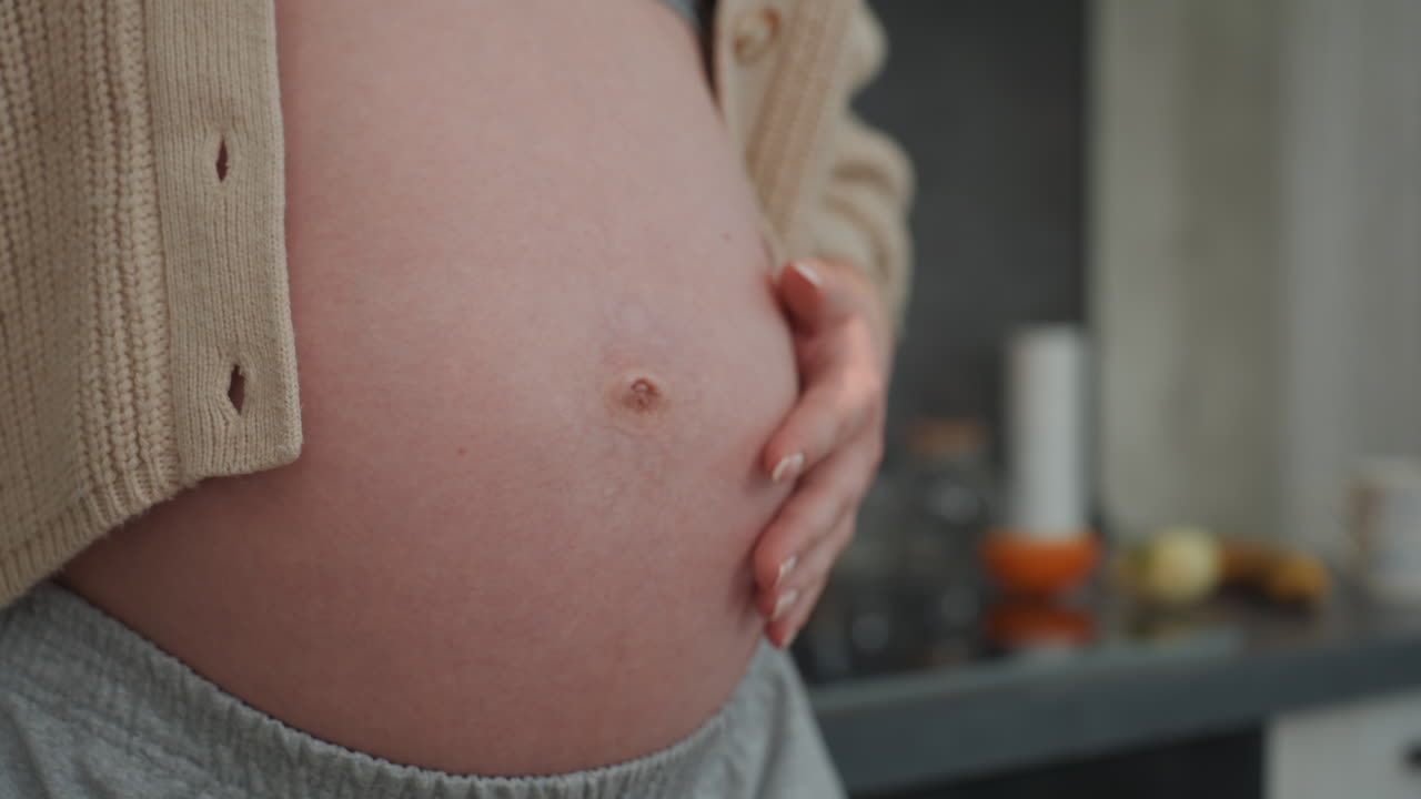 Primo piano di una donna bianca incinta che accarezza la pancia con mani delicate, con ombelico e texture della pelle visibili. Lo sfondo è un salotto soffuso. Un automassaggio prenatale calmante, con un cardigan comodo e una posa rilassata, crea un'atmosfera intima.