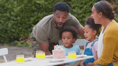 Familia-Del-Ejército-Celebrando-El-Cumpleaños-Del-Niño-En-El-Jardín-De-Casa-Apagando-Velas-En-El-Pastel