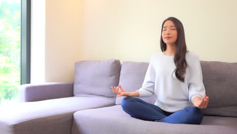 asian young woman meditating on her sofa in the living room near a window