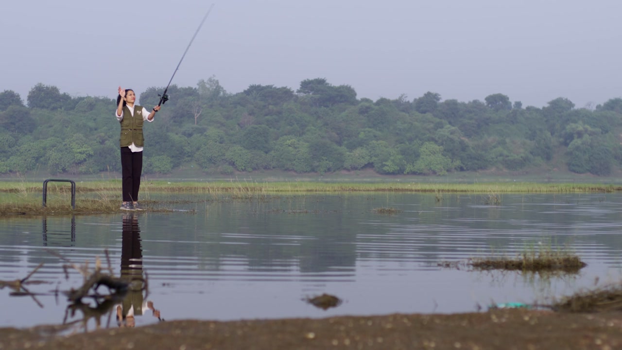A South Asian girl wearing an outdoor vest fishes near wetlands, holding her rod while standing in water
