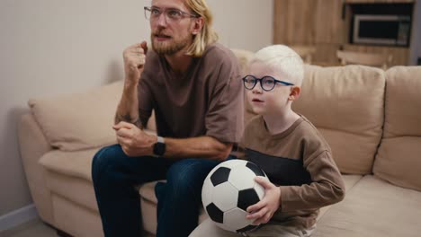 Ein-Glücklicher-Kleiner-Albino-Junge-Mit-Weißem-Haar-Und-Blauer-Brille-Springt-Auf-Und-Wirft-Einen-Fußball,-Während-Er-Mit-Seinem-Blonden-Vater-Mit-Bart-Und-Brille-Zu-Hause-Auf-Dem-Sofa-Ein-Fußballspiel-Ansieht