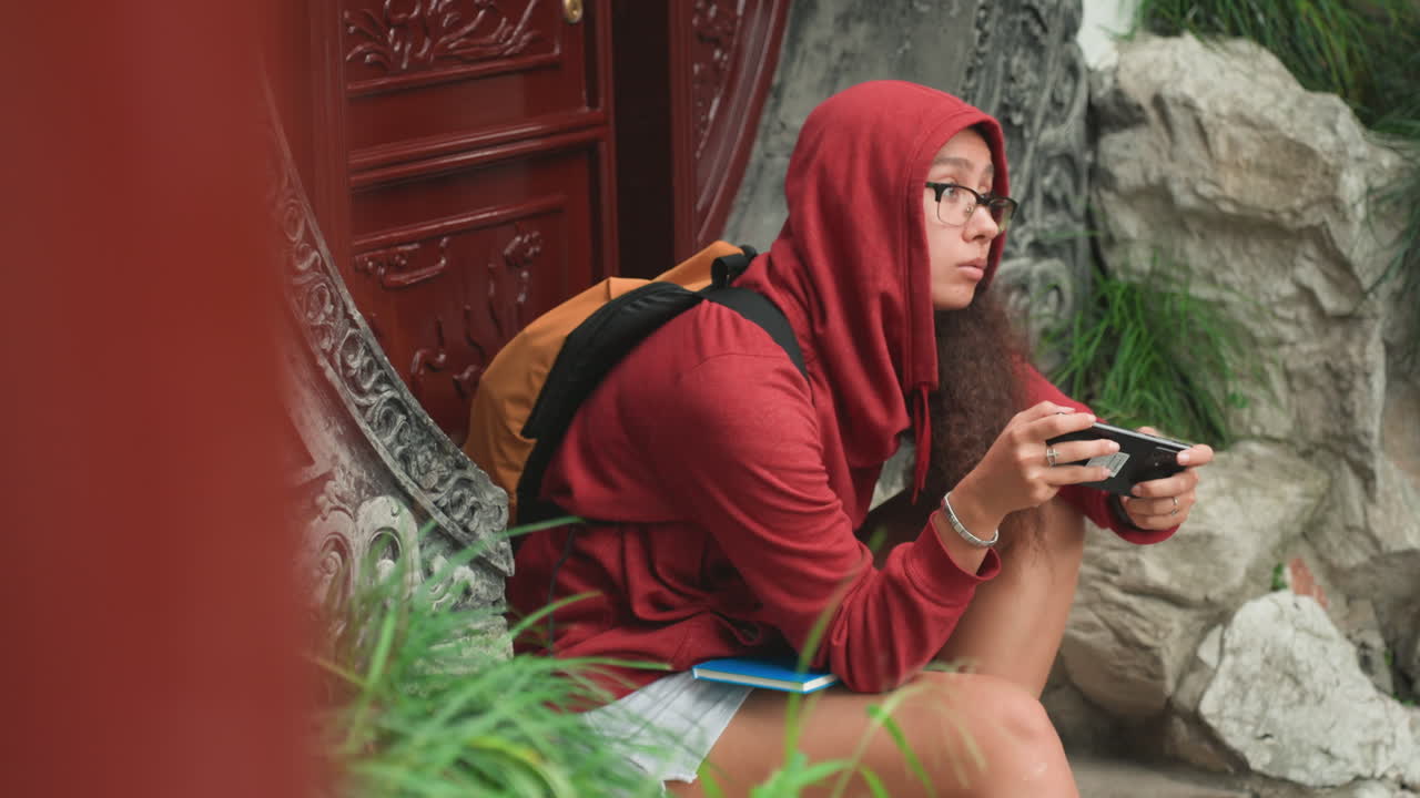 Pengunjung wanita berhijab menggunakan perangkat di samping pintu masuk artistik. Wanita berpenutup kepala menjelajahi sekitar dan memeriksa ponselnya di dekat gerbang batu dekoratif di tengah taman