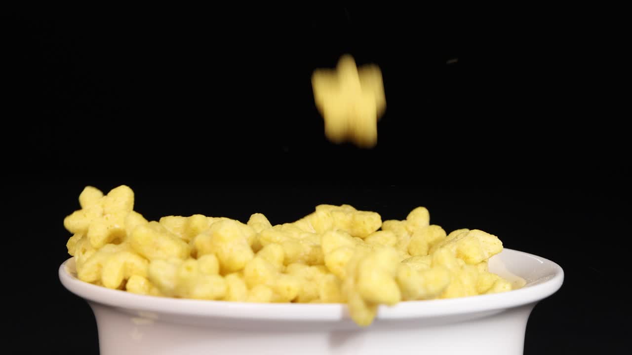 Yellow cereal pieces fall into a white bowl against a black background, captured in slow motion for a dynamic visual effect