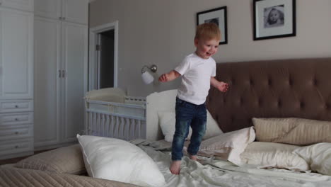 a boy in a white t-shirt jumps on the bed and laughs at the sight of soap bubbles in the parents' bedroom in slow motion