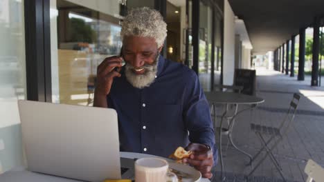 african american senior man talking on smartphone while having breakfast sitting outdoors at cafe