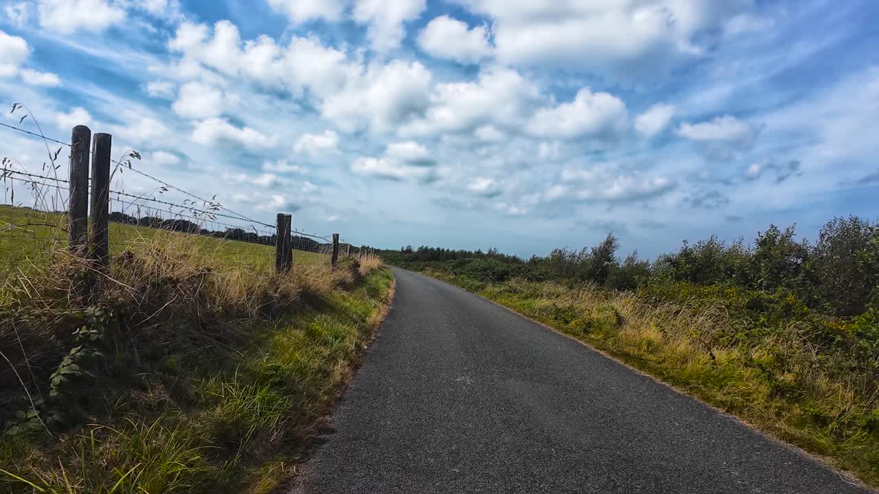 Slow Moving POV on Rural Road with Fence and Fields Passing By in Natural Environment with Bright Blue Cloud Sky. Example of British Countryside Cycling