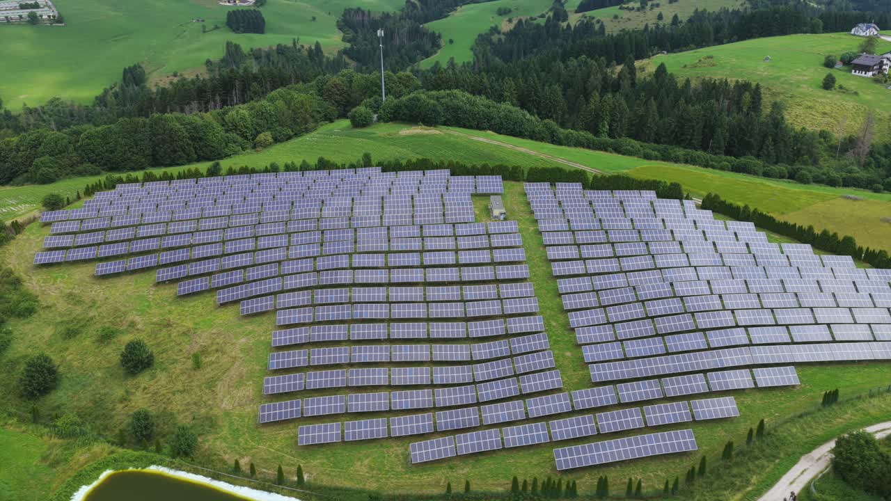 Drone orbit of a hillside solar farm: sweeping rows of photovoltaic panels across green pastures, ringed by forest and rolling hills. Clean energy, sustainability, renewable power footage