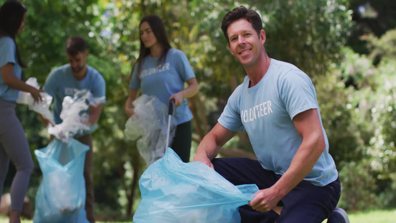 hombre caucásico sonriente y un grupo diverso de amigos poniendo basura en bolsas de basura azules en el parque