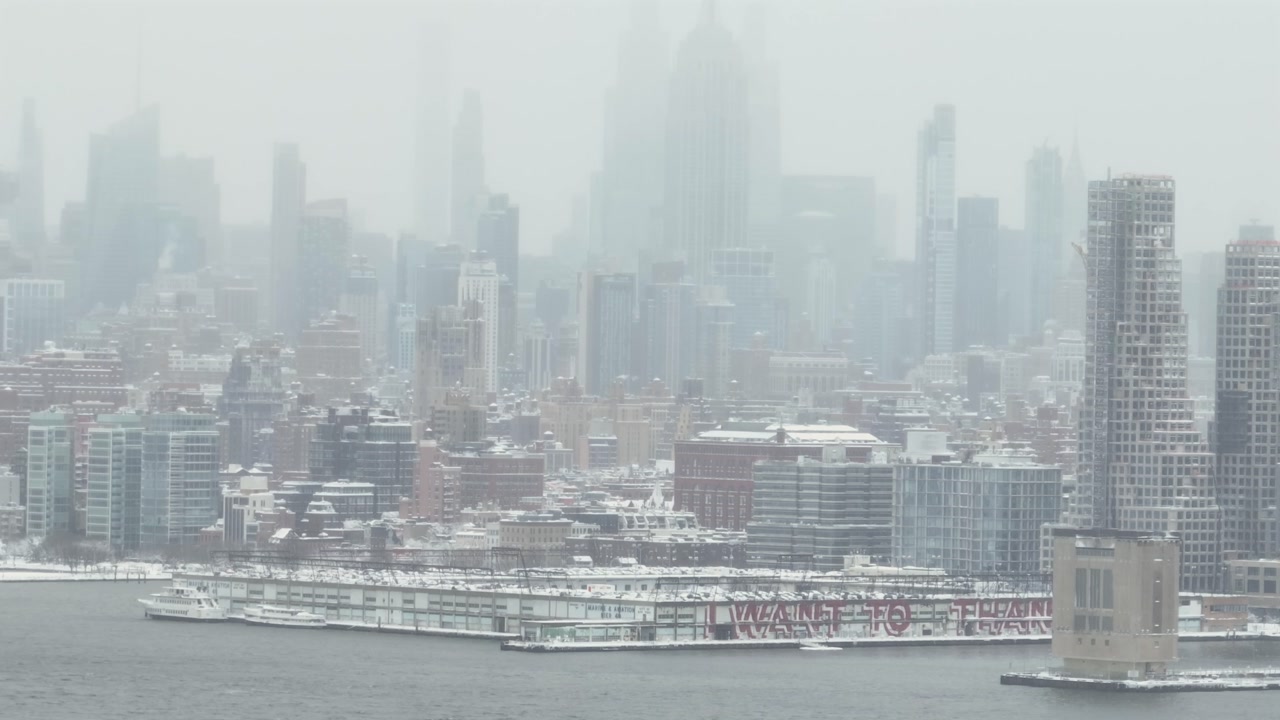 Wide aerial view of Jersey City skyline in winter, with dense fog and low clouds partially obscuring skyscrapers along the waterfront, creating a muted, cold and atmospheric urban scene.