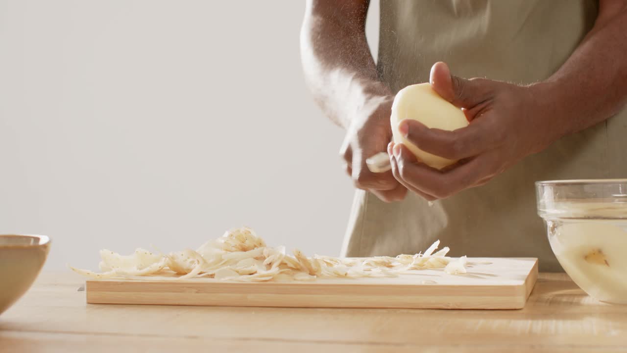 Un uomo con il grembiule inizia a preparare un pasto in cucina, sbucciando una cipolla intera con un pelapatate su un tagliere.