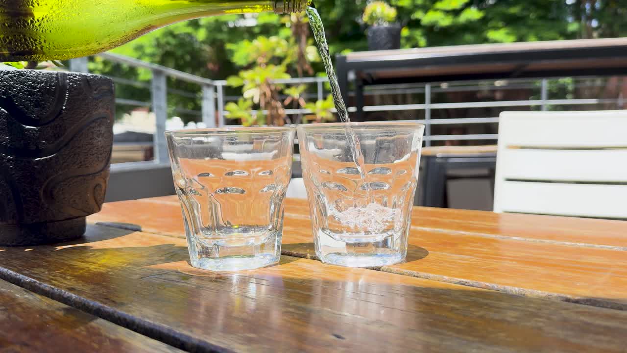 Water is poured into glasses on a sunlit outdoor table, creating a refreshing and vibrant atmosphere