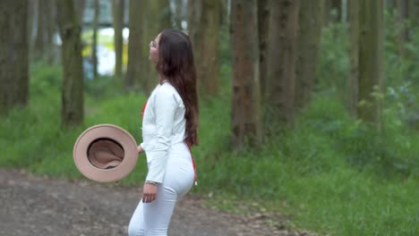 a woman in white attire gracefully walking through the forest, gently removing her hat amidst towering trees, embodying a peaceful escape into nature