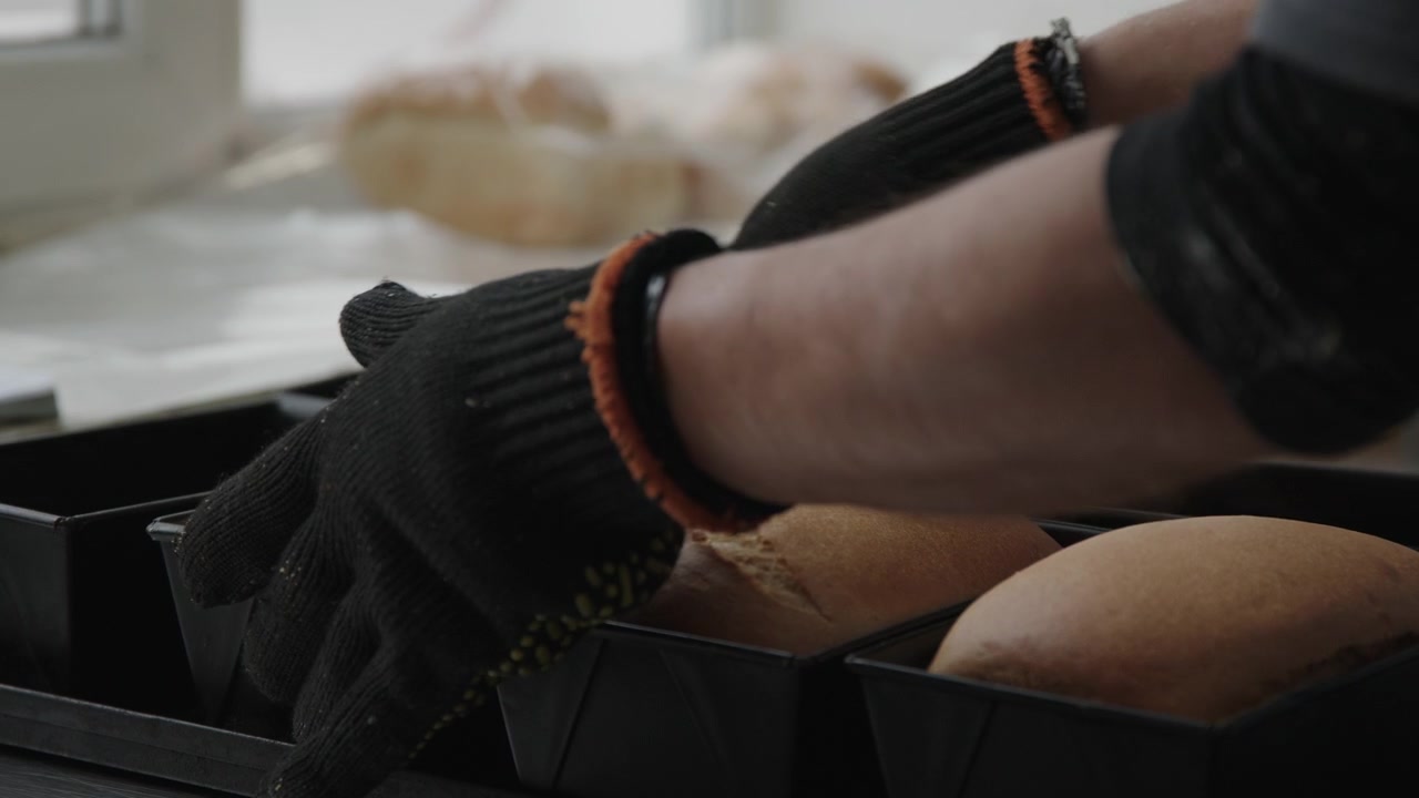 Una persona mette il pane in teglie rettangolari su un piano di lavoro della cucina. Il pane è caldo e appena sfornato. La cucina è luminosa con luce naturale.