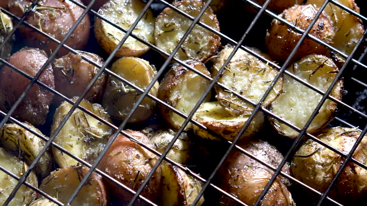 붉은 불씨가 아래에서 빛나는 뜨거운 그릴에 있는 와이어 바구니에 있는 갈색 허브가 있는 새 감자를 똑바로 내려다보고 패닝하는 카메라