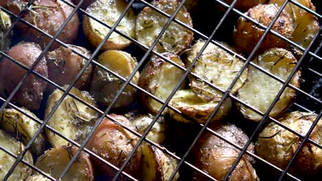 camera looking straight down and panning across browned herbed new potatoes in a wire basket on a hot grill with red embers glowing beneath