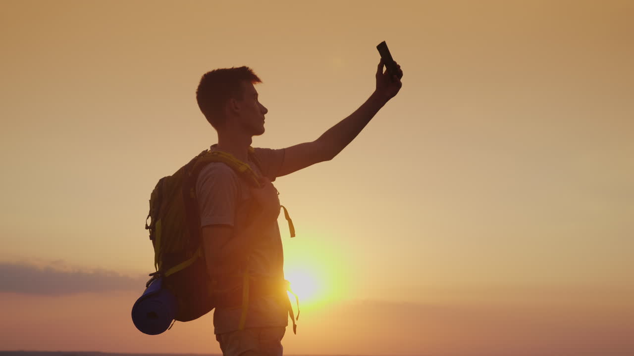 Un adolescente con uno zaino si scatta un selfie con il telefono sullo sfondo del tramonto. Turismo e viaggi.