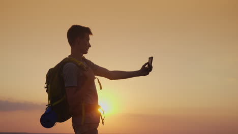 A-Teenager-With-A-Backpack-Pictures-Himself-With-A-Phone-Against-The-Setting-Sun-Tourism-And-Travel-