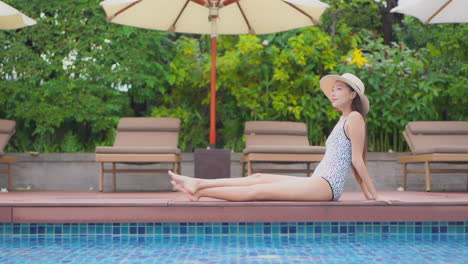 asian model woman sitting stretching her legs at the edge of the swimming pool daytime in thailand resort, side view