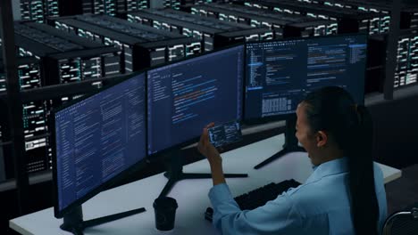 Side View Of Happy Asian Woman Developer Looking At Code On Mobile Phone While Write Code With Multiple Computer Screens In Data Center