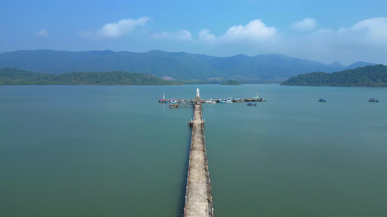 Long Walkway pier with a lighthouse reaching into blue ocean with mountains under clear sunny sky. overflight flyover fly push forward drone