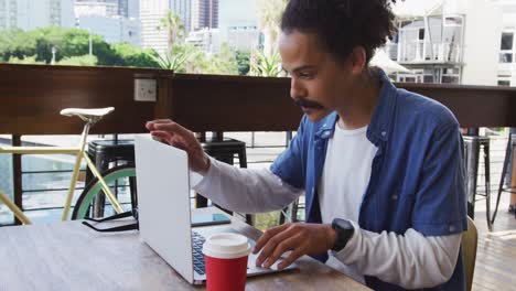 mixed race man w ith moustache sitting at table outside cafe using laptop