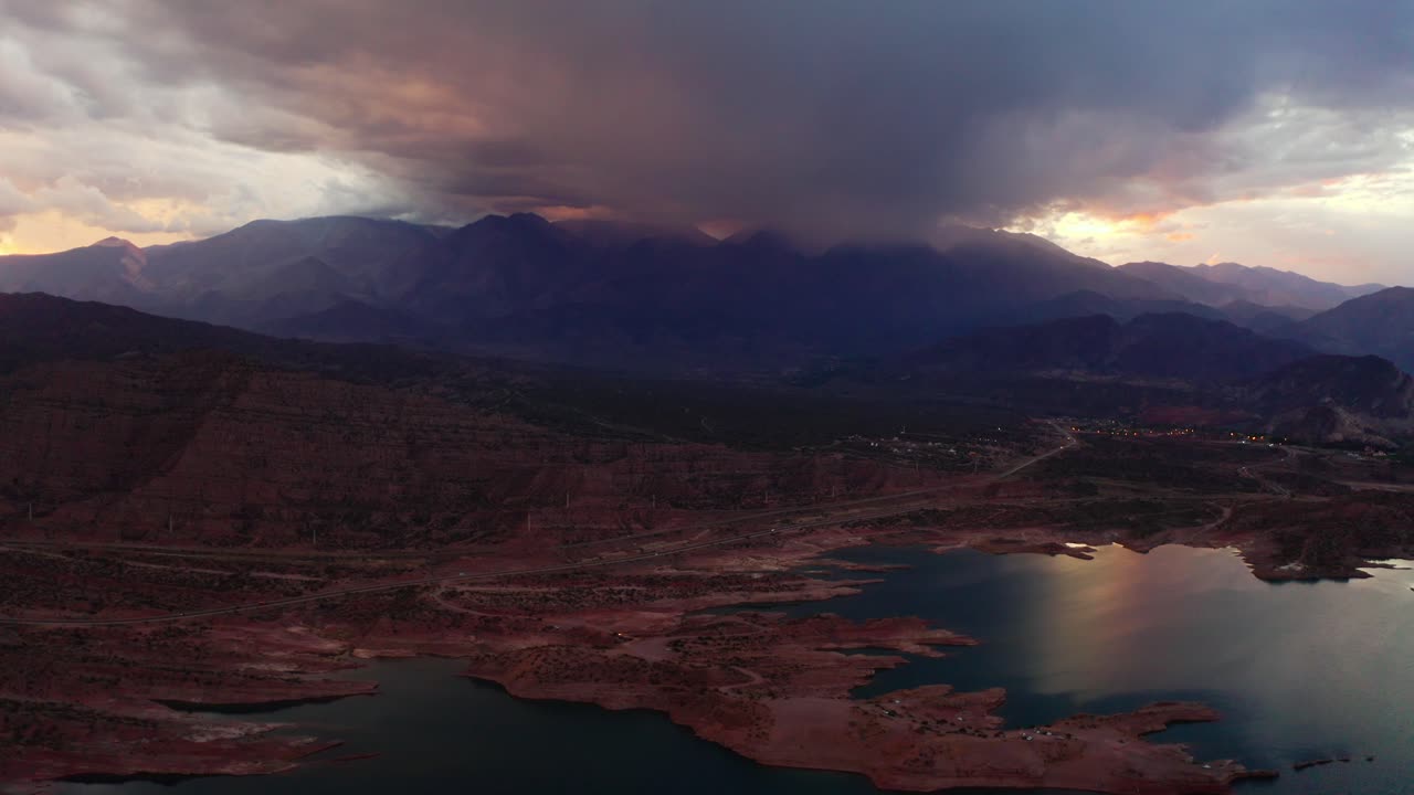 Lightning storm across Potrerillos reservoir surrounded by Andean mountains at dusk, Mendoza Argentina, aerial pullback