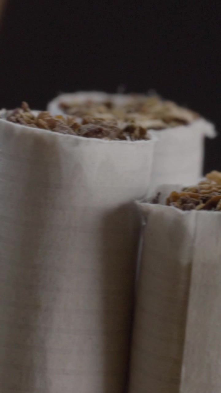 Close-up panning shot moves across cigarettes, revealing white paper wrappers, loose brown tobacco filling, cylindrical forms, and fine surface texture against dark background.