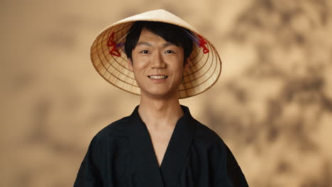 Young-Asian-man-in-black-traditional-costume-and-conus-hat-smiling-at-camera