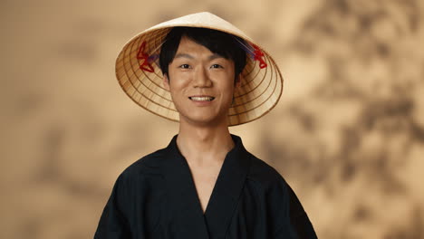 young asian man in black traditional costume and conus hat smiling at camera