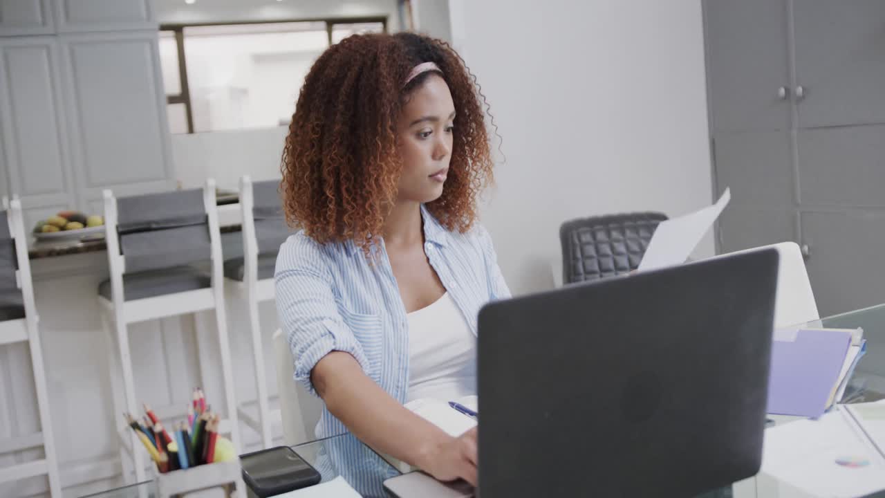 Busy biracial woman working on laptop and inspecting documents at home, in slow motion