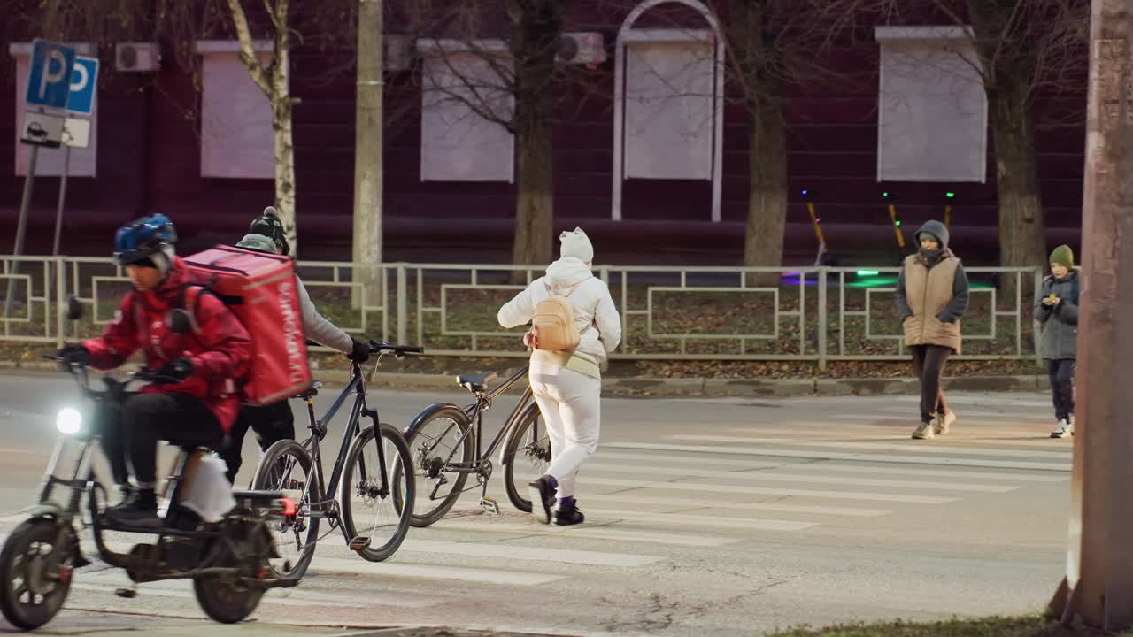 Urban evening scene showing people in warm winter clothes walking bicycles across striped pedestrian crossing while a delivery rider approaches on electric bike under street lighting