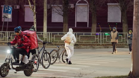 urban evening scene showing people in warm winter clothes walking bicycles across striped pedestrian crossing while a delivery rider approaches on electric bike under street lighting