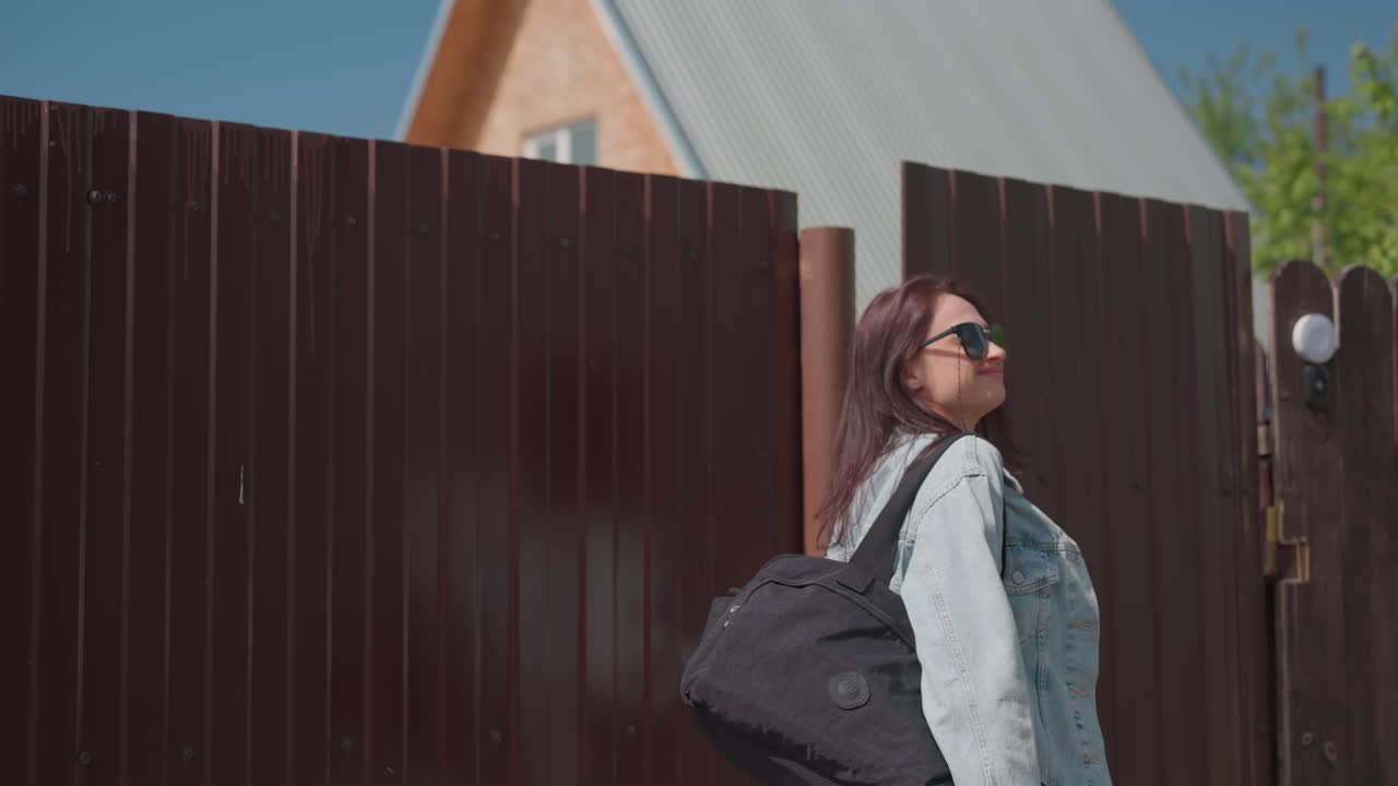 Smiling young woman in white top opens metal gate to enter residential yard on sunny day, preparing to walk through entrance as friend follows behind