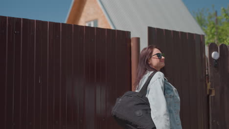 smiling young woman in white top opens metal gate to enter residential yard on sunny day, preparing to walk through entrance as friend follows behind