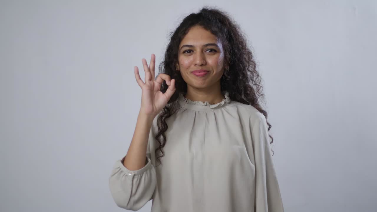 A South Asian young woman with curly hair smiles, making an 'okay' gesture with her hand against a gray background