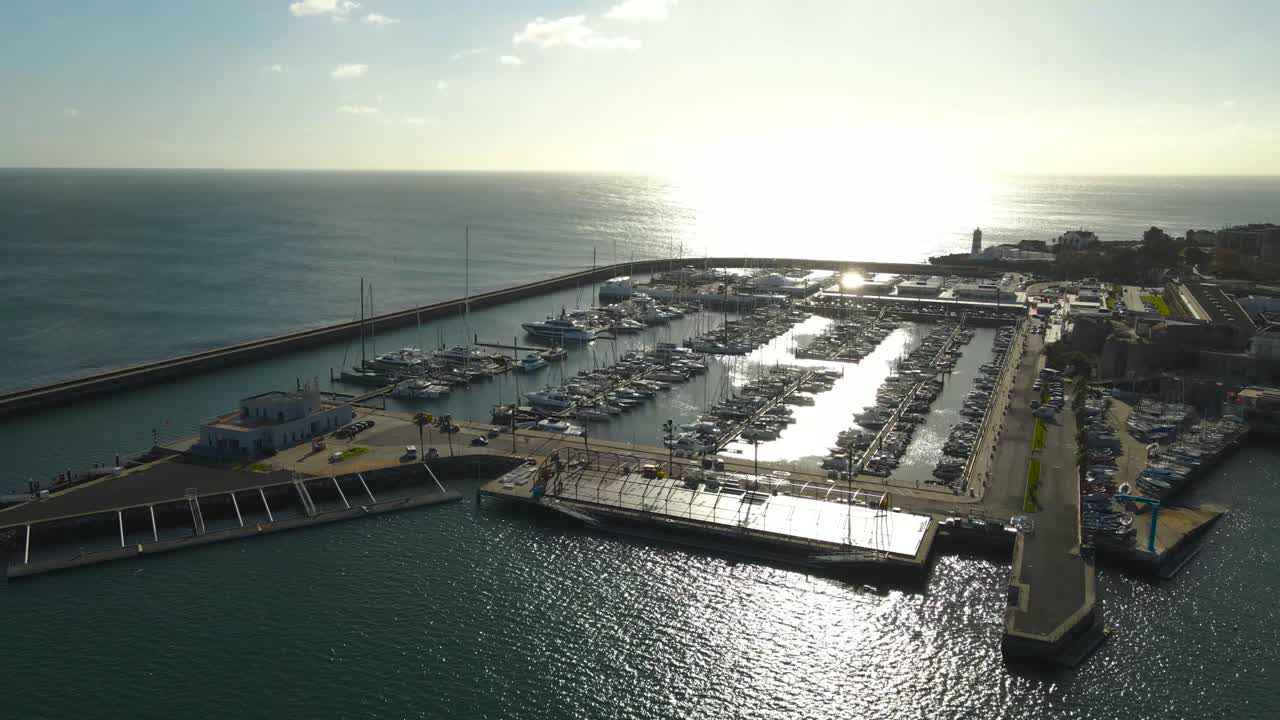 Órbita suave de dron sobre el puerto deportivo de Estoril en Cascais con yates de lujo, la costa del Océano Atlántico y la brillante luz del sol de verano reflejándose en el agua