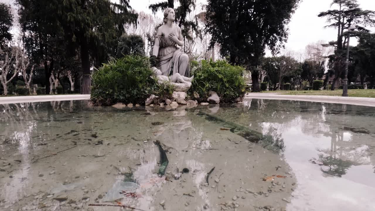 Sculpture surrounded by murky water in a tranquil garden near ancient trees on a cloudy day