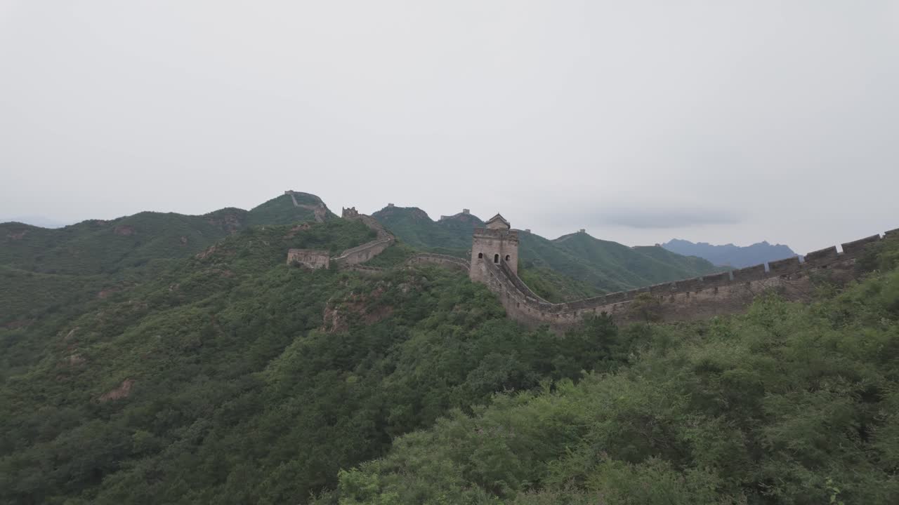 Eine Weitwinkelaufnahme der Chinesischen Mauer, die sich durch grüne Bergkämme unter einem bedeckten Himmel schlängelt. Perfekt für Reisedokumentationen und historische Inhalte.