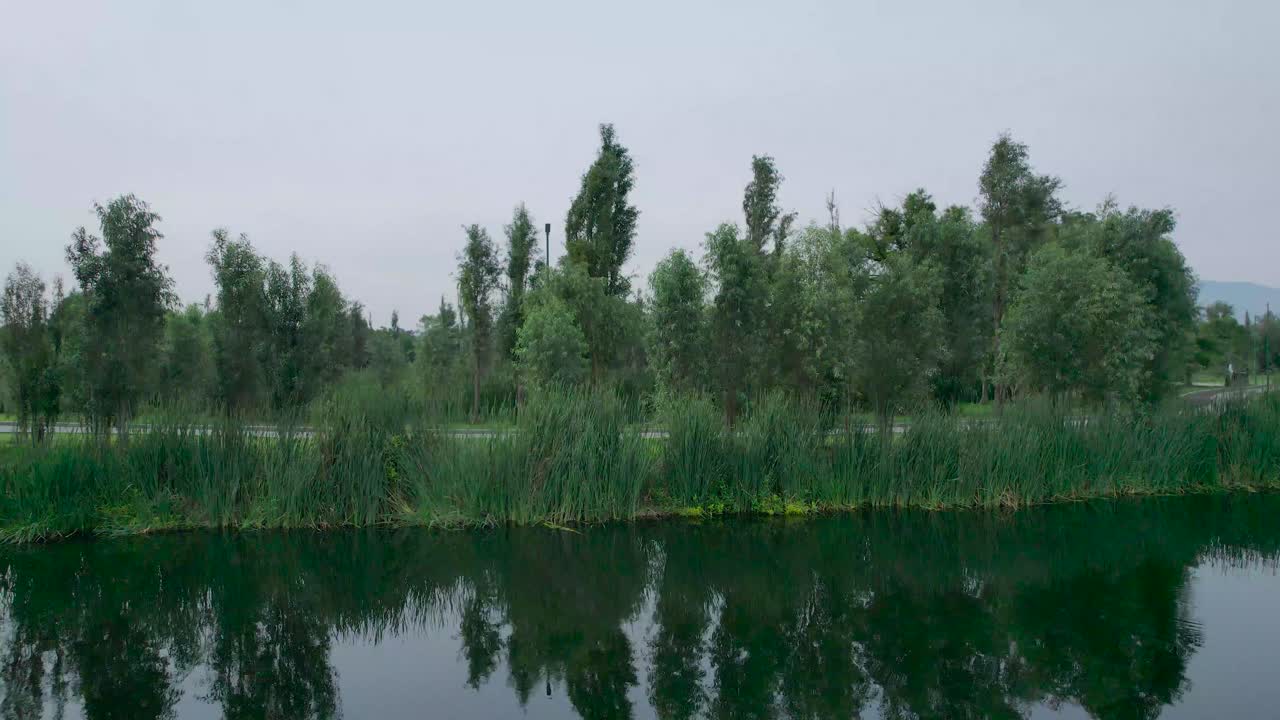Pull-right to left above a lake, with view of a training trail in natural area in Mexico City, with view of some birds and trees. Footage finish in front of mexican boats named trajineras