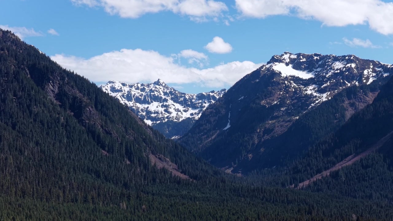 Passo Snoqualmie con cime rocciose innevate e una tranquilla valle profondamente boscosa, fitti pendii sempreverdi che si ergono su entrambi i lati sotto un cielo azzurro brillante con nuvole sparse. Washington, USA. Ampia ripresa aerea.