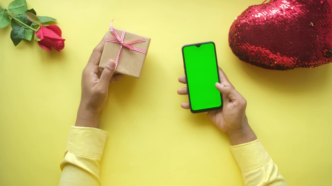 mano sosteniendo teléfono inteligente con pantalla verde y regalo en fondo amarillo