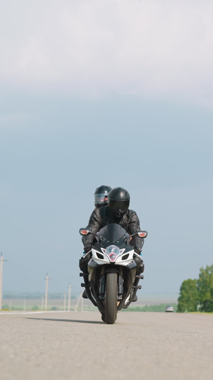 People with helmets ride fast motorcycle along long road against green tree windbreak under sky with white clouds at countryside slow motion