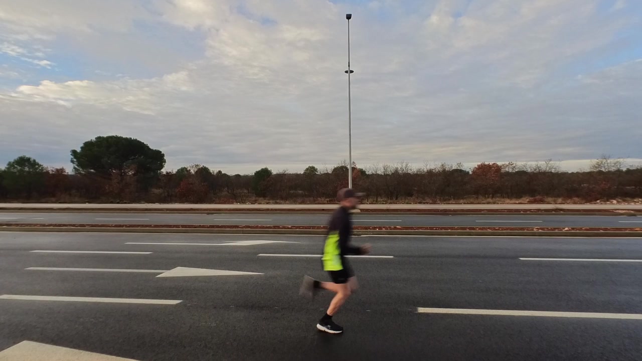 Side view of an athletic man jogging across a wide, empty asphalt road during a cloudy sunrise. Concept of endurance, steady pace, and outdoor fitness.