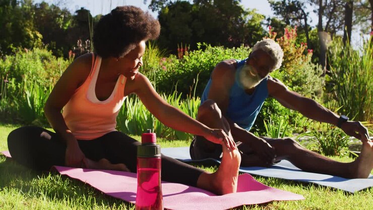 African american senior couple exercising outdoors sitting stretching in sunny garden
